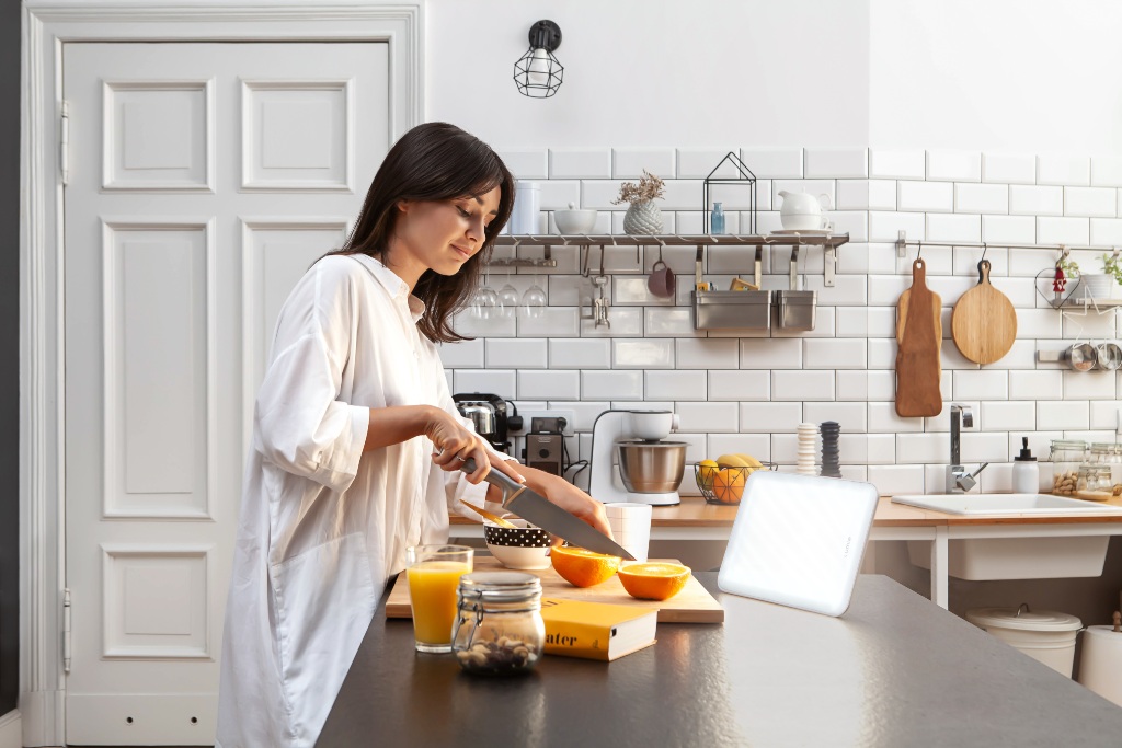 Woman cutting fruit in kitchen in front of a Lumie Vitamin L SAD light