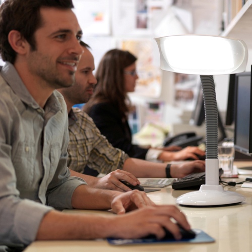 Man working next to colleagues at computers, with Lumie Desklamp SAD light, a type of bright light therapy device.