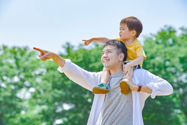Man with a little boy on his shoulders outdoors. They are both pointing to something out of picture. The sun is shining, and they have some skin exposed, so would be making Vitamin D if the UV index was 3 or higher.